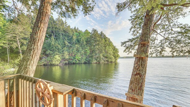 A wooden deck with a steering wheel, overlooking a calm lake surrounded by dense forest. - Peaceful Lakefront Maine Cabin w/ Sunset Views (Vassalboro)