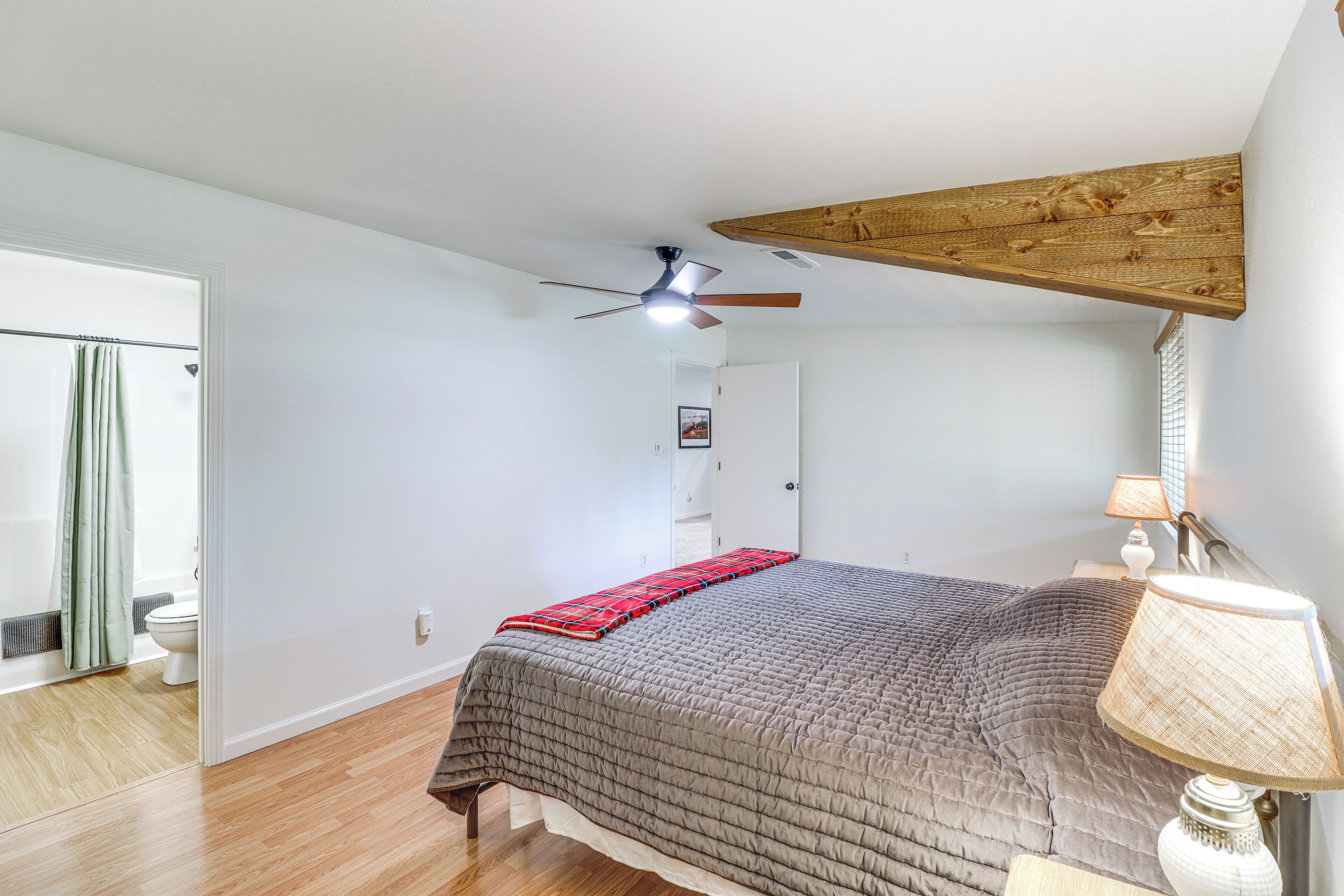 Un dormitorio con cama, ventilador de techo, viga de madera, mesita de noche con lámpara y baño visible a través de una puerta abierta.
