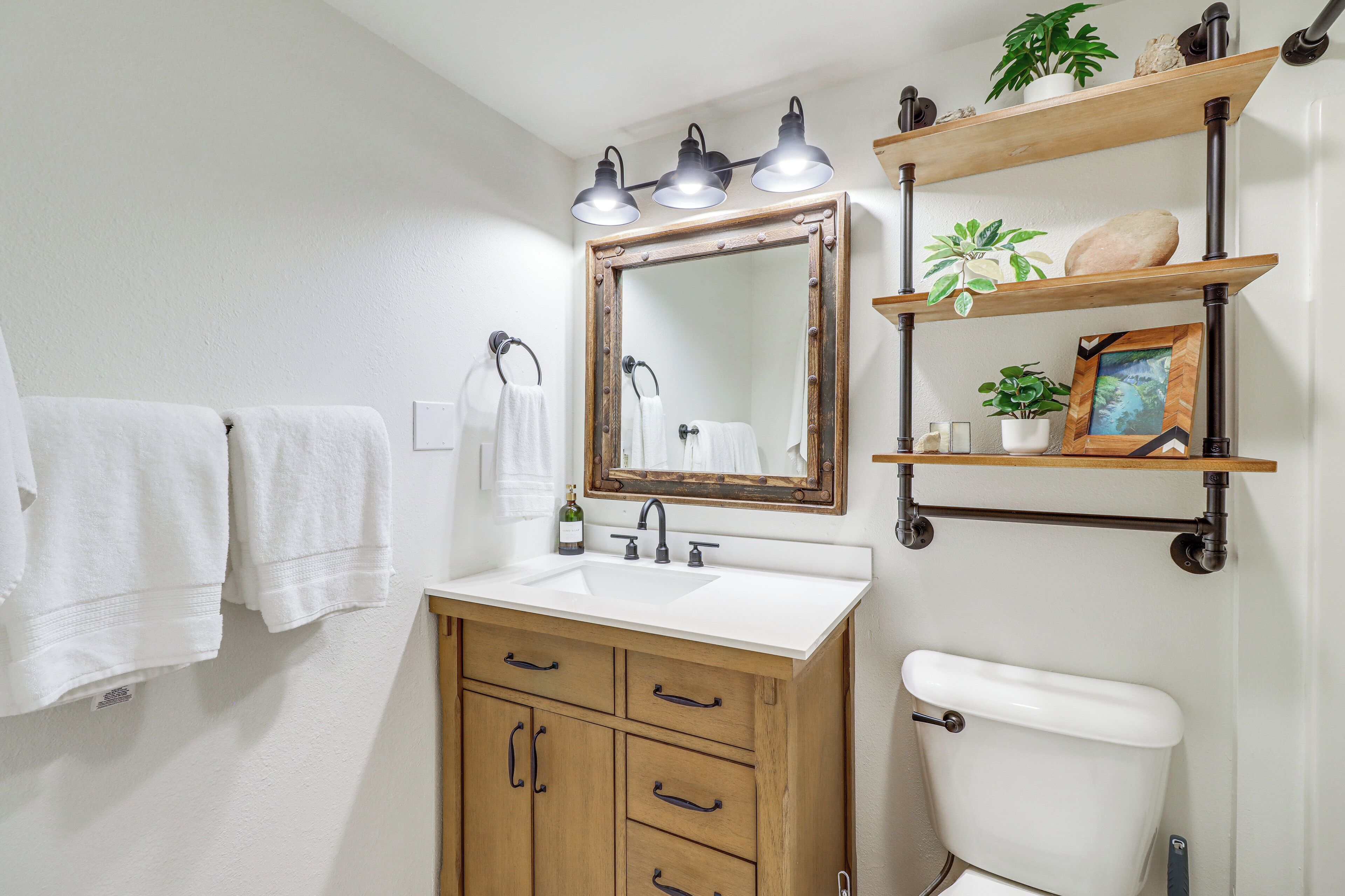 Un baño con tocador de madera, un lavabo blanco, un espejo y un inodoro. Hay toallas colgadas en la pared y estantes con plantas y un marco de fotos.