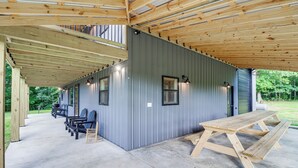 A covered outdoor area with a picnic table and benches, a metal building with windows, and a wooden ceiling structure.