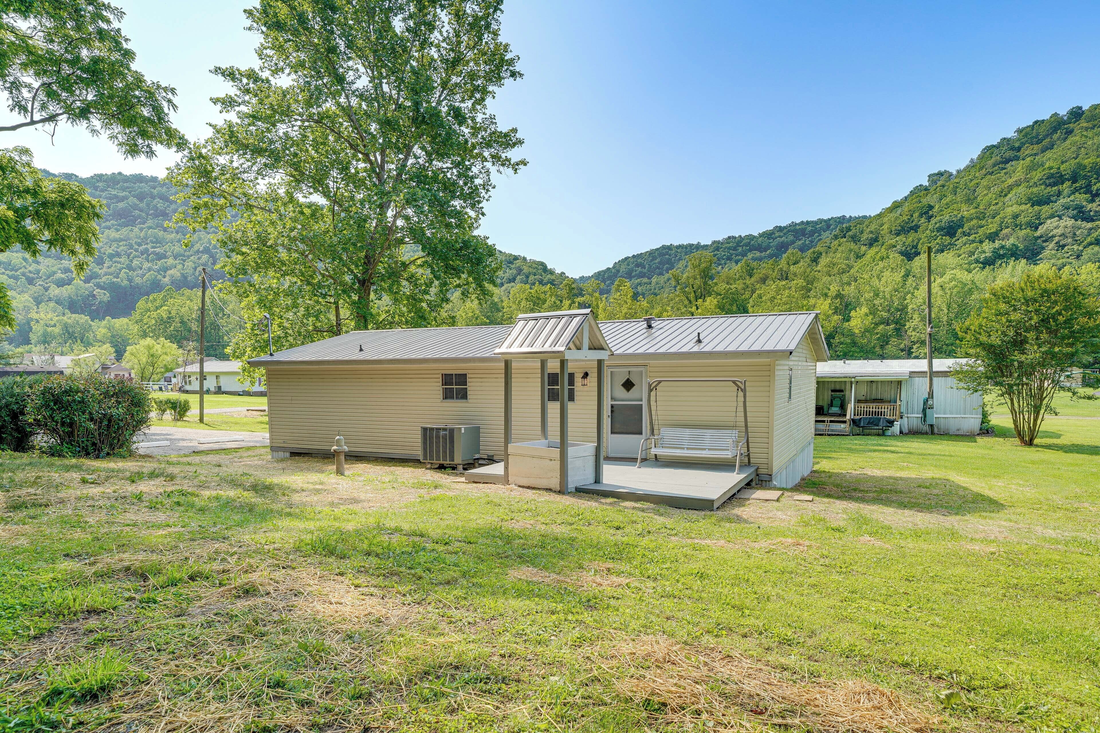 A small, single-story building with a covered porch and a small deck, surrounded by a grassy yard and trees, set against a backdrop of hills.