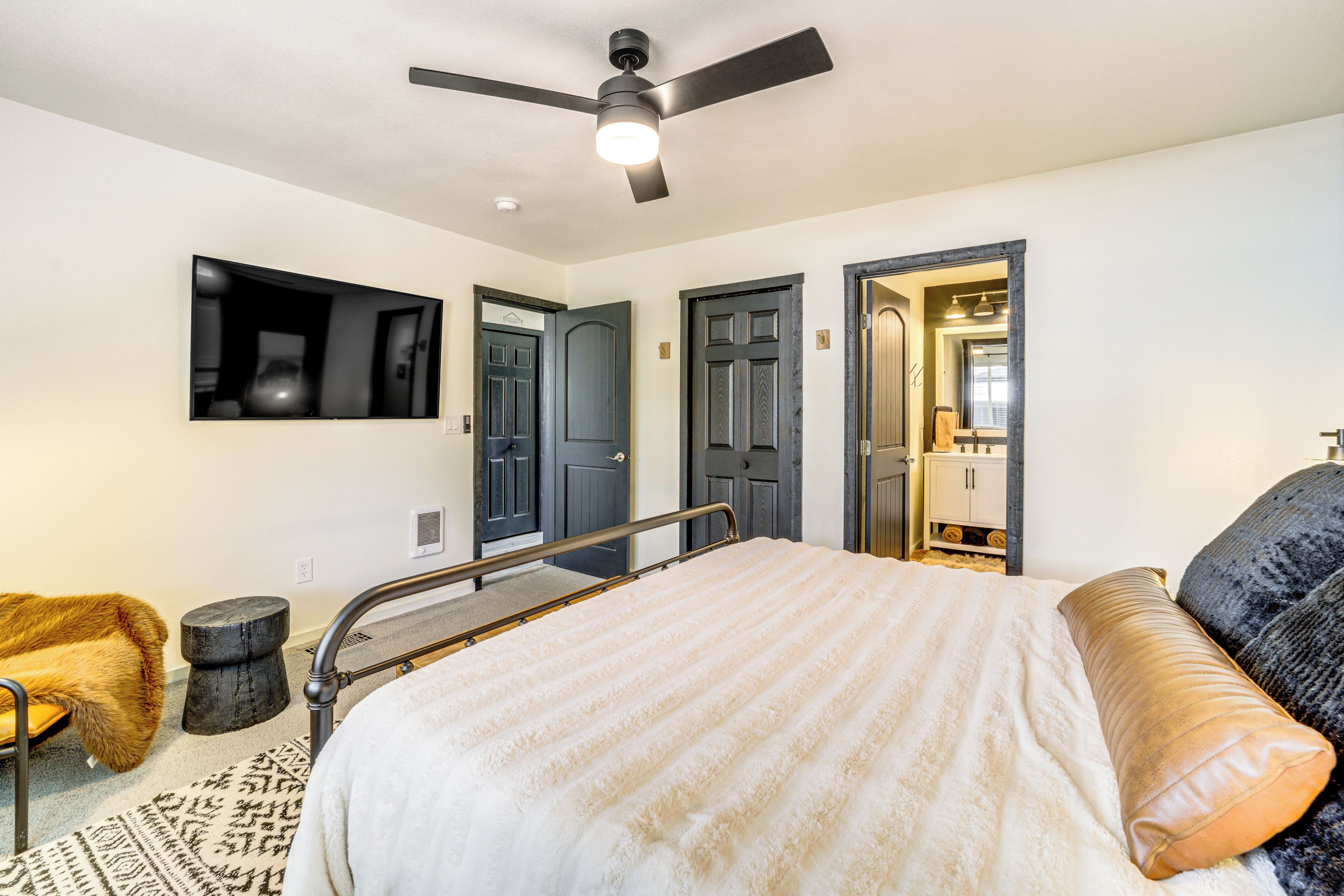A bedroom with a bed, a ceiling fan, a television mounted on the wall, and a door leading to another room.