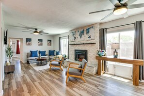A spacious living room with hardwood flooring, a fireplace, a ceiling fan, and a large window with curtains. - Newly Renovated Albuquerque Retreat w/ Pool Table (Albuquerque)