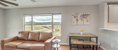 A modern living room with a leather sofa, a round coffee table, and a dining table with chairs. There is a large window providing a view of a landscape.