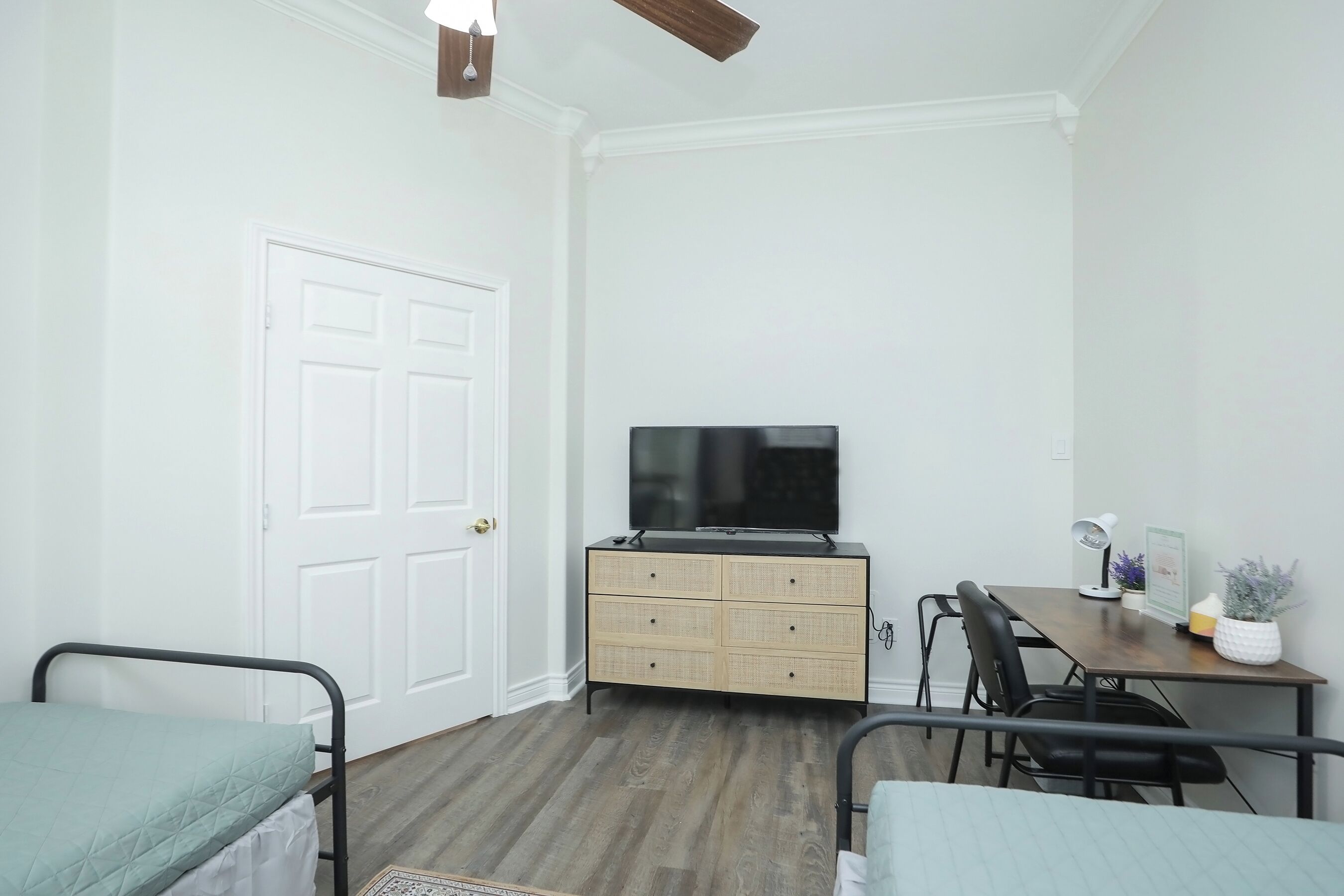 A bedroom with a bed, a dresser, a desk with a chair, a television, and a ceiling fan.