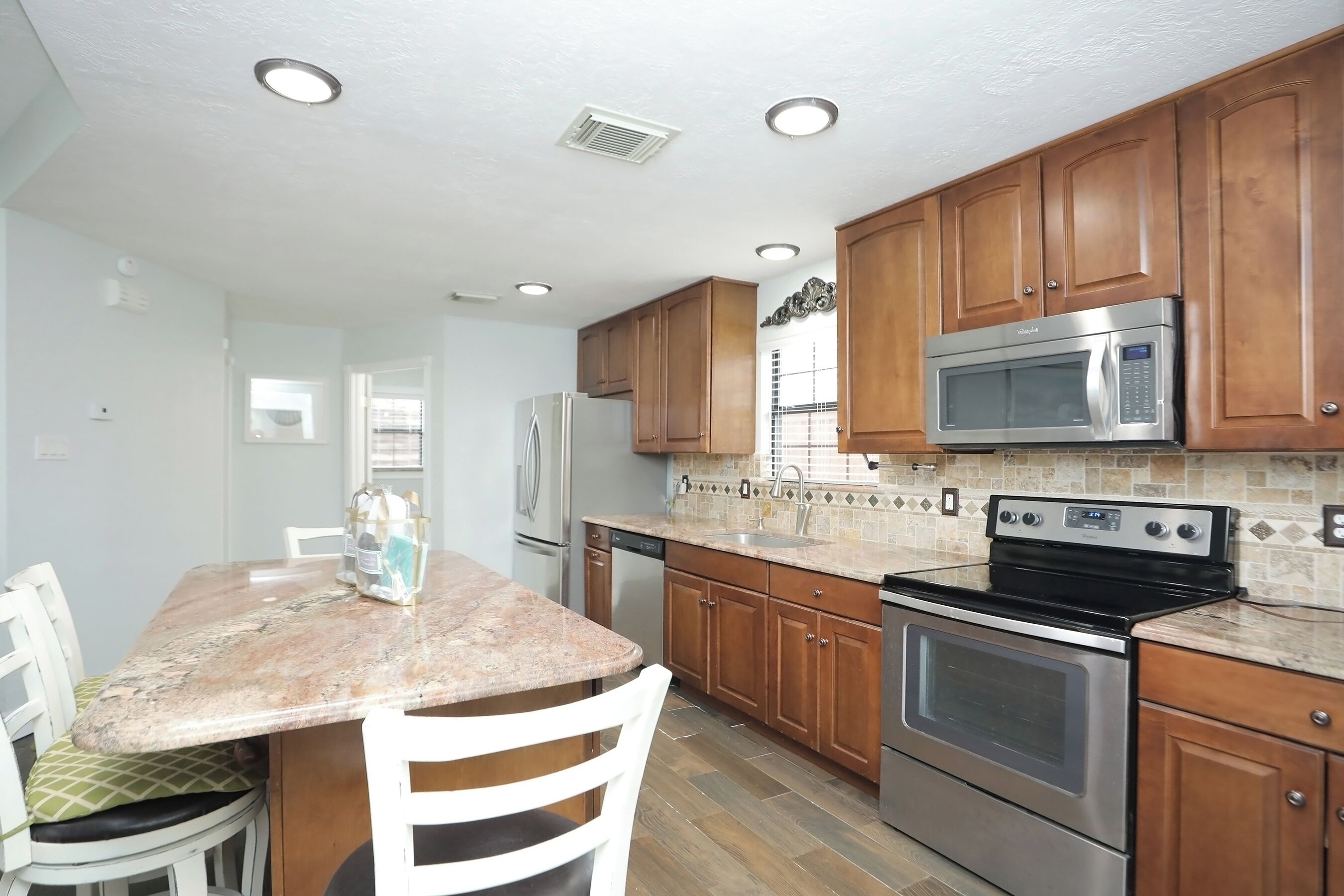 A kitchen with wooden cabinets, a granite countertop, stainless steel appliances, and a dining area with white chairs.