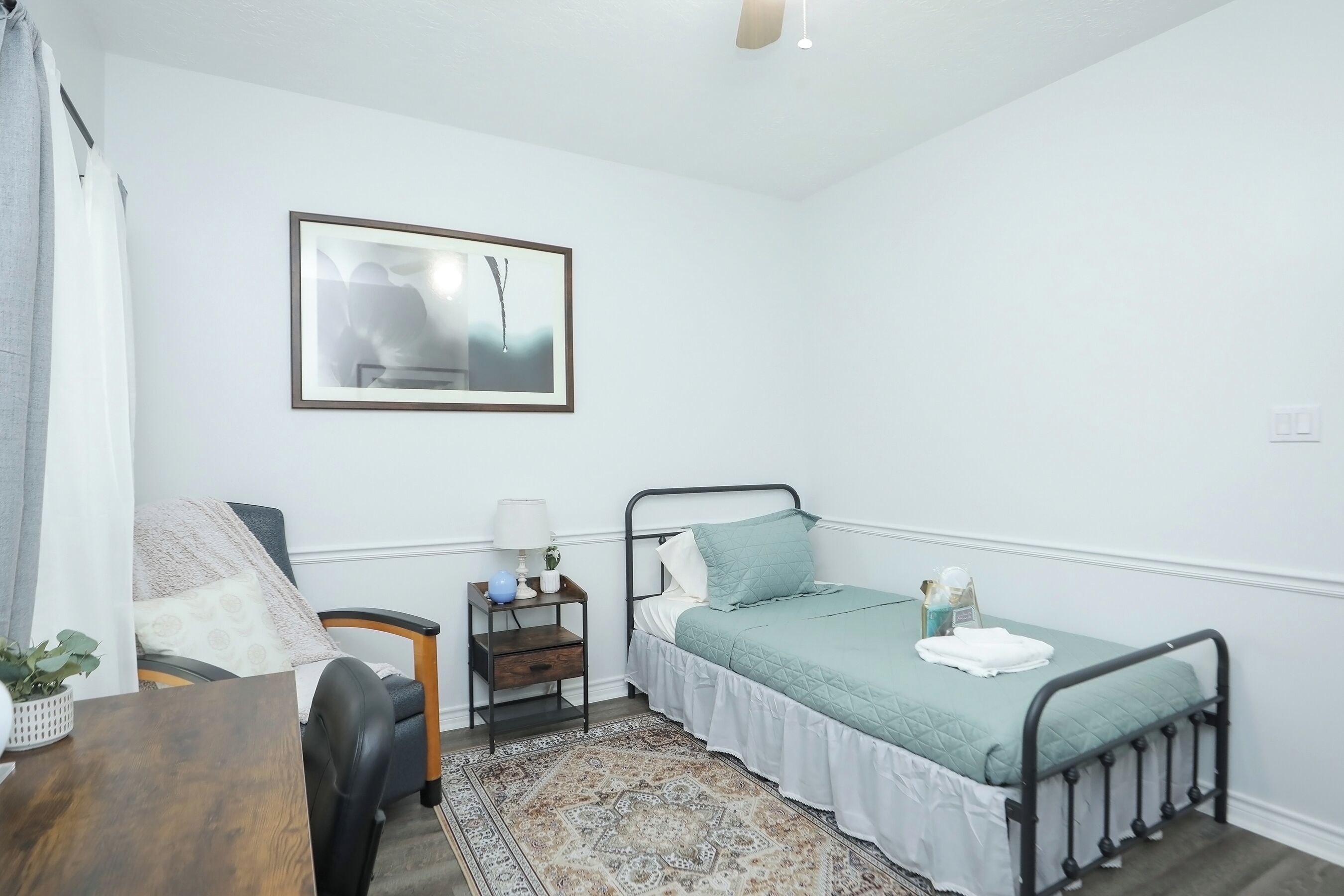 A small, neatly arranged bedroom with a single bed, a chair, a nightstand with a lamp, and a framed picture on the wall.