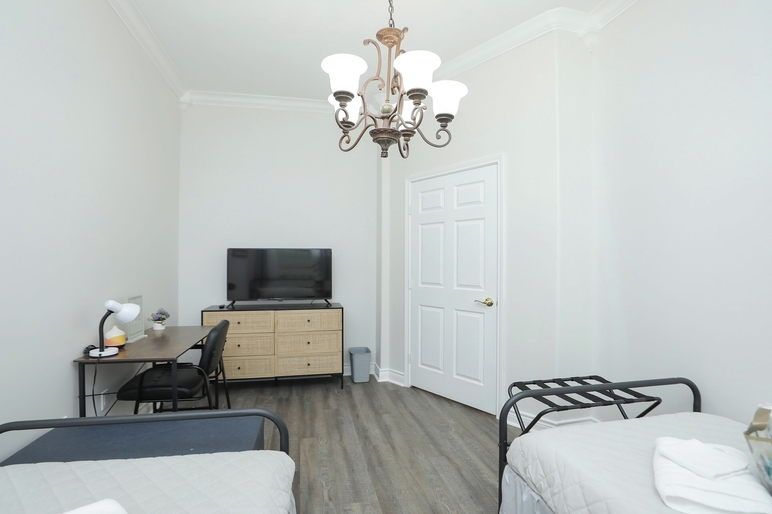 A bedroom with two beds, a desk with a chair, a television, and a chandelier.