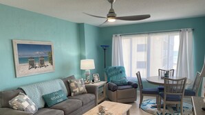Un salon avec un mur de couleur turquoise, un ventilateur de plafond, une table à manger avec des chaises, un canapé avec des coussins et un tableau encadré accroché au mur.