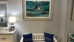 Une chambre avec un tableau représentant une tortue de mer, encadré, une table de chevet avec une lampe et un berceau avec deux oreillers.