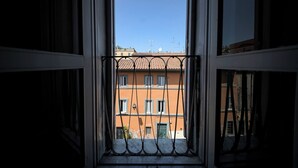 Une vue à travers une fenêtre ornée d’une grille métallique, donnant sur un bâtiment avec un toit recouvert de tuiles et de nombreuses fenêtres.