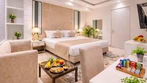 A modern hotel room with a large bed, bedside tables, a seating area with a coffee table, and a small kitchenette.