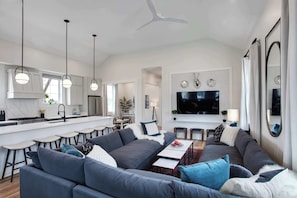A modern living room with a grey sofa, a dining table, and a kitchen area with bar stools. - Luxe 19BR Suit | Private Backyard with Pool (New Orleans)