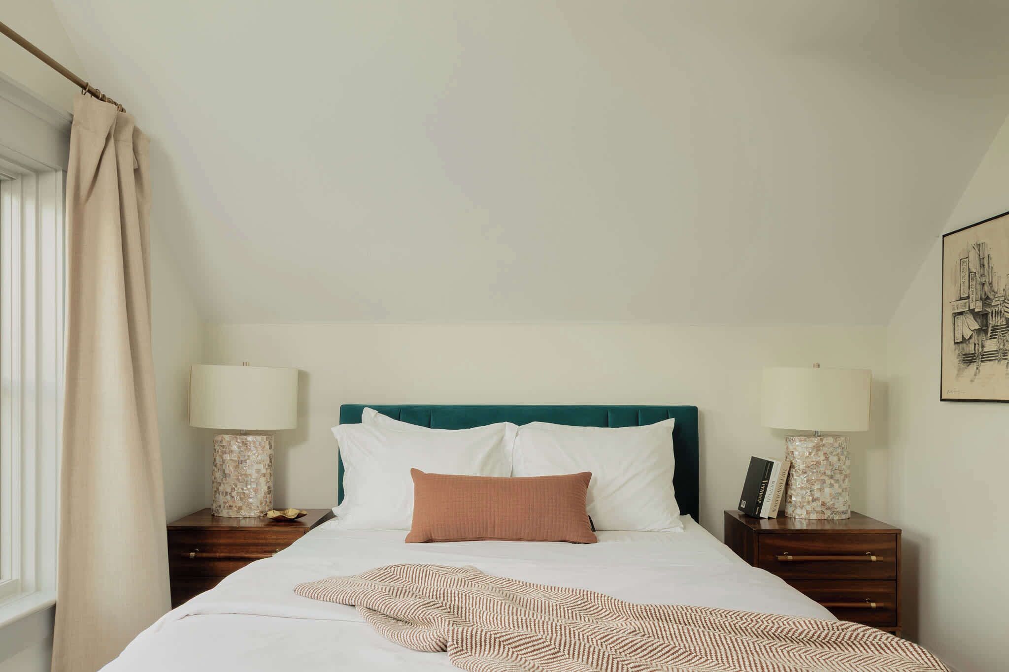 A neatly made bed with white and brown bedding, two bedside tables with lamps, and a window with curtains.