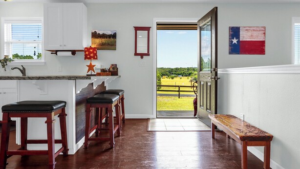 A kitchen with wooden stools, a bench, and a view of a green field through a door. - Clare House I Couples, Stargazing (San Marcos)