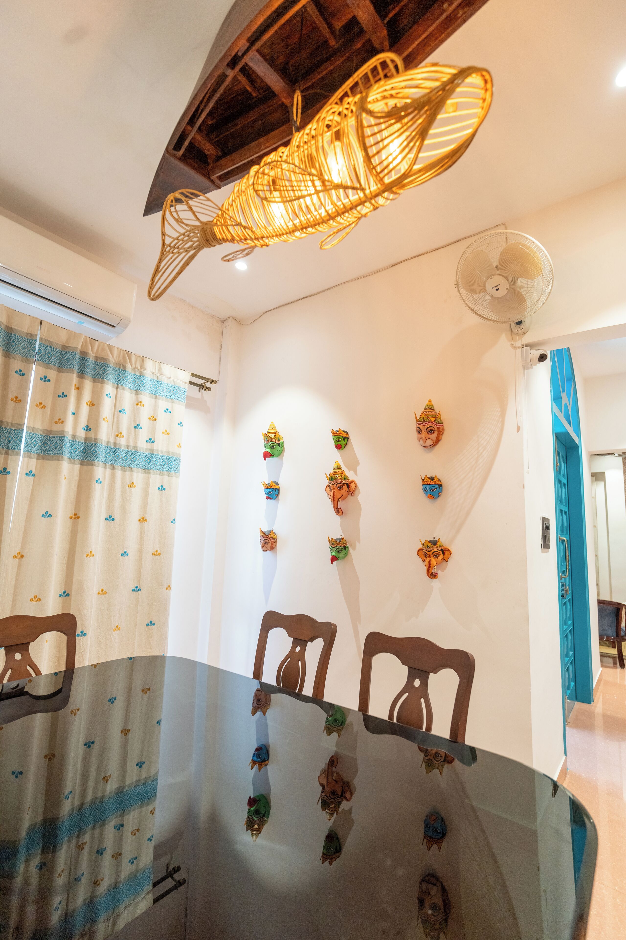 Een eetgedeelte met een ronde glazen tafel, stoelen, een plafondventilator en decoratieve wandkunst.