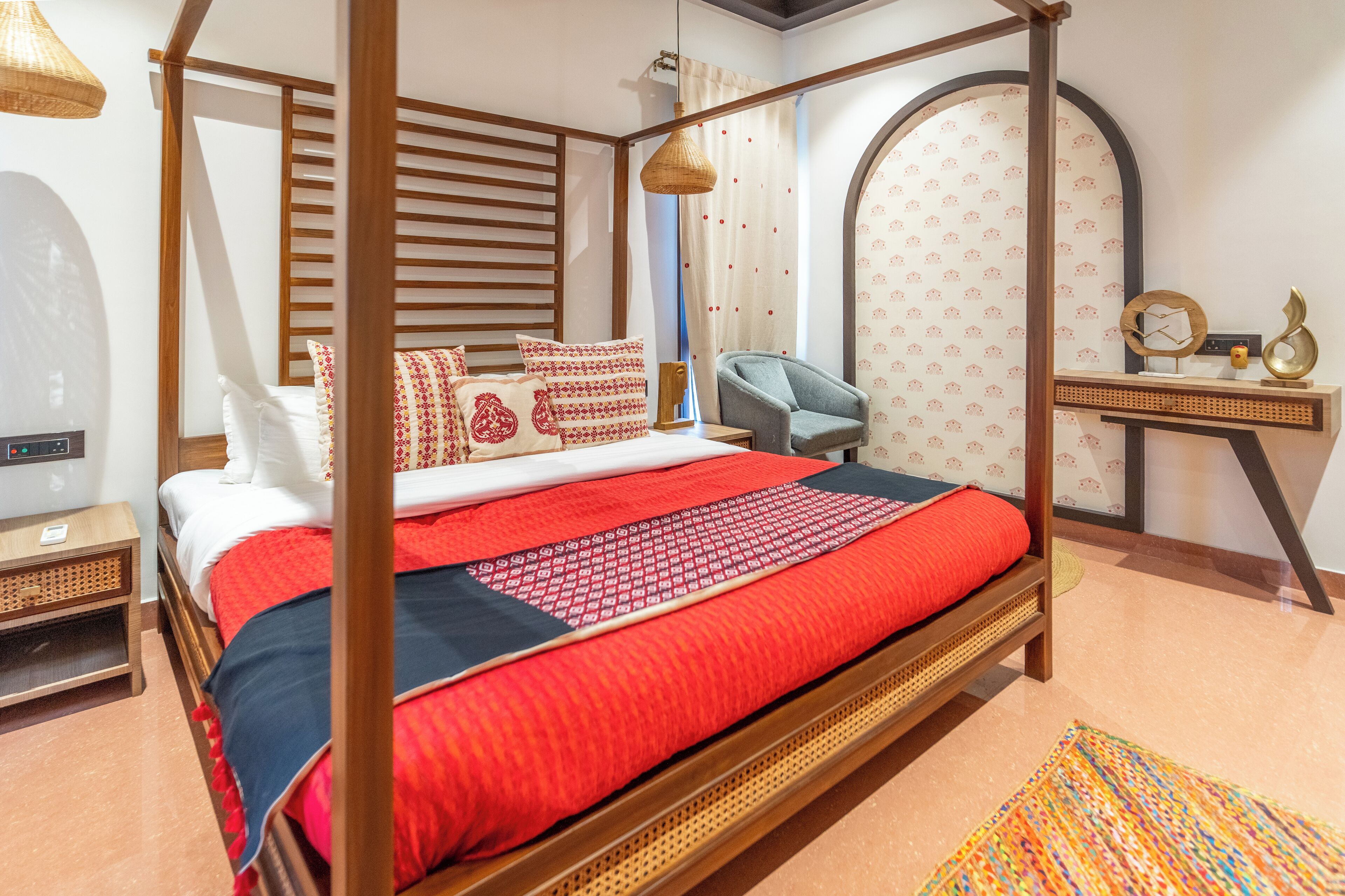 Una cama con dosel con un edredón rojo y negro, un sillón gris, una mesita con un espejo y objetos decorativos, y una pared con diseño.