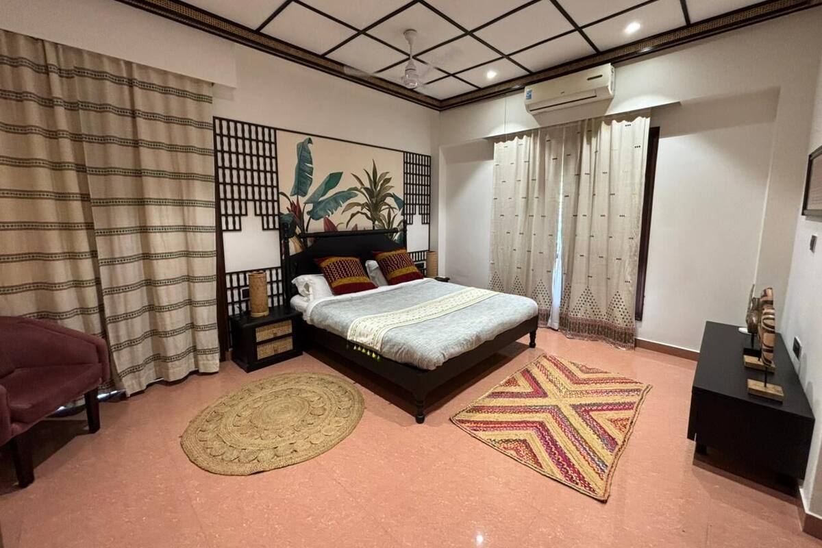 Quarto com cama, mesa de cabeceira, ar-condicionado fixado na parede, janela com cortinas e um objeto decorativo na parede.