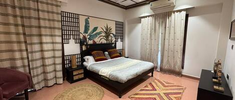 A bedroom with a bed, a nightstand, a wall-mounted air conditioner, a window with curtains, and a decorative wall piece.