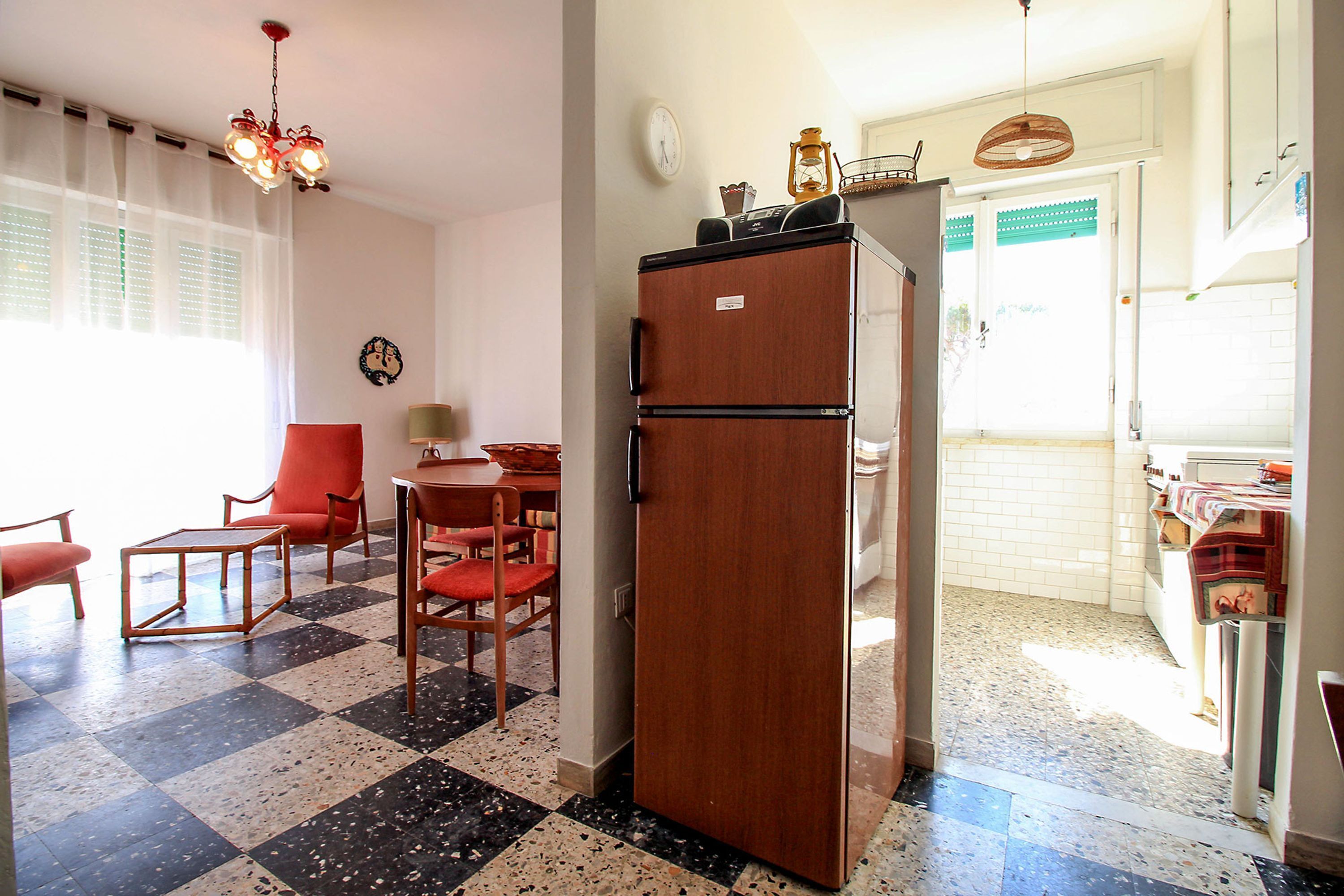 A room with a checkered floor, a wooden refrigerator, a dining table with chairs, and a kitchen area with a sink and cabinets.