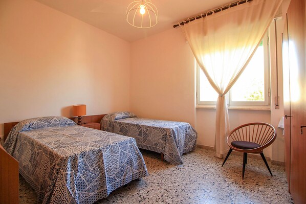 A room with two beds, a chair, a lamp, and a window with curtains. - Appartamento Anna 100 m dal Mare (Castiglione della Pescaia)