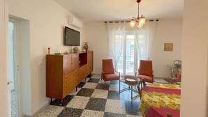 A room with a checkered floor, a wooden cabinet, two chairs, a table with a floral tablecloth, a television, and a ceiling light fixture.