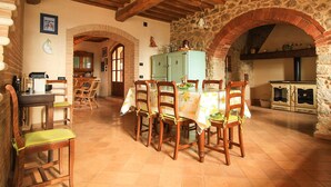 A rustic dining area with a wooden table and chairs, a stone wall, and a kitchen in the background. - Villa Este con Piscina (Civitella Paganico)