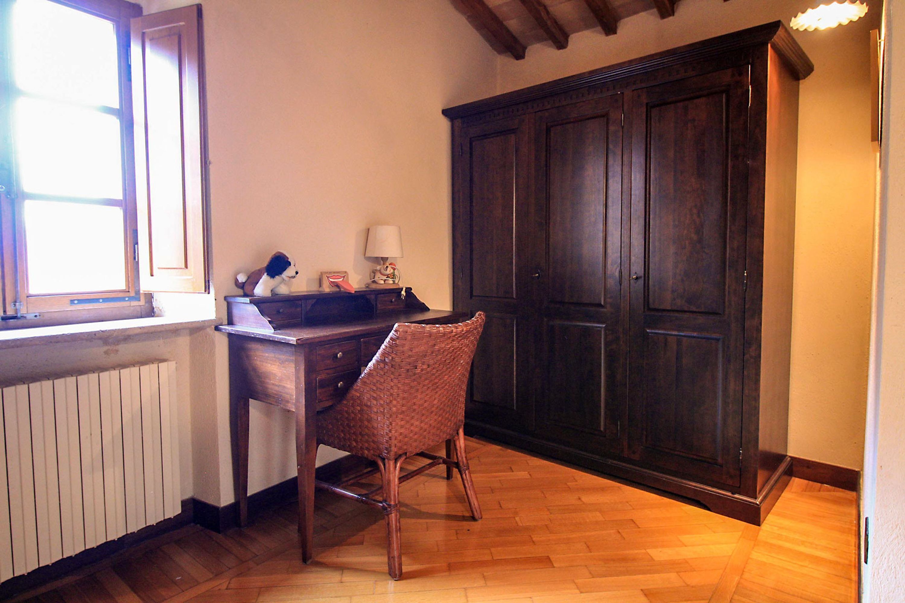 Ein Zimmer mit einem Holztisch, einem Korbstuhl, einem dunklen Holzschrank und einem Fenster mit Jalousien.