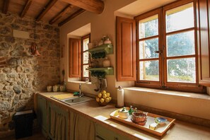 Une cuisine rustique avec des murs en pierre, des poutres en bois, un évier et une fenêtre donnant sur un paysage verdoyant.