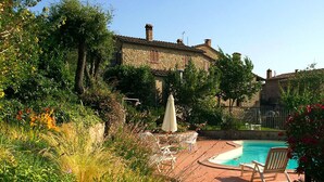 A pool area with lounge chairs and a table, surrounded by lush greenery and stone buildings. - Villa Este con Piscina (Civitella Paganico)