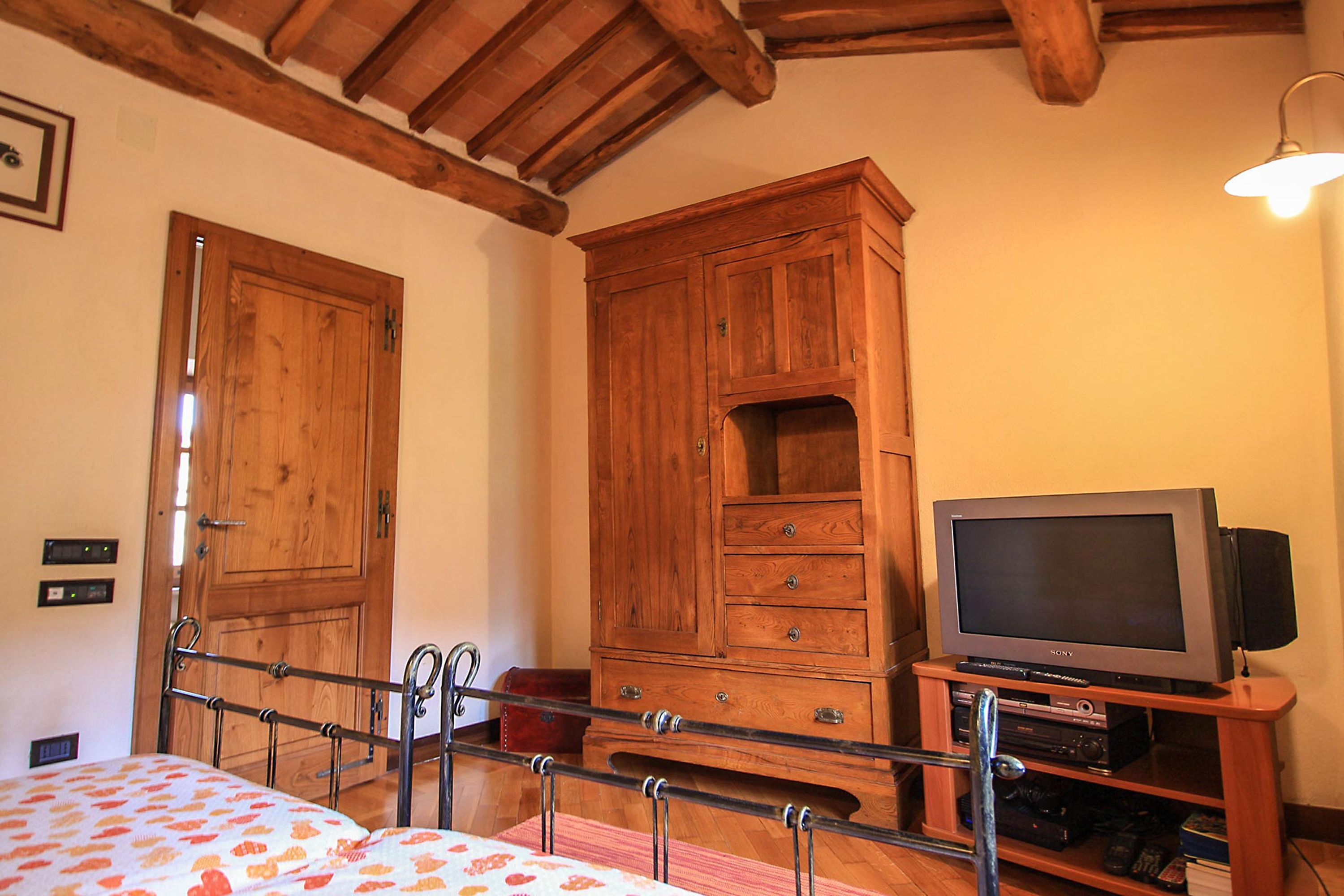 Une chambre avec une armoire en bois, un lit recouvert d’une couvre-lit à motifs floraux, un téléviseur posé sur un meuble en bois et un plafond avec des poutres apparentes.