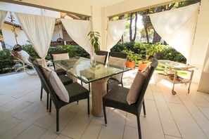 Une table à plateau en verre, entourée de chaises en osier dotées de coussins, est placée sur une terrasse carrelée agrémentée de plantes en pot et offrant une vue sur la verdure.