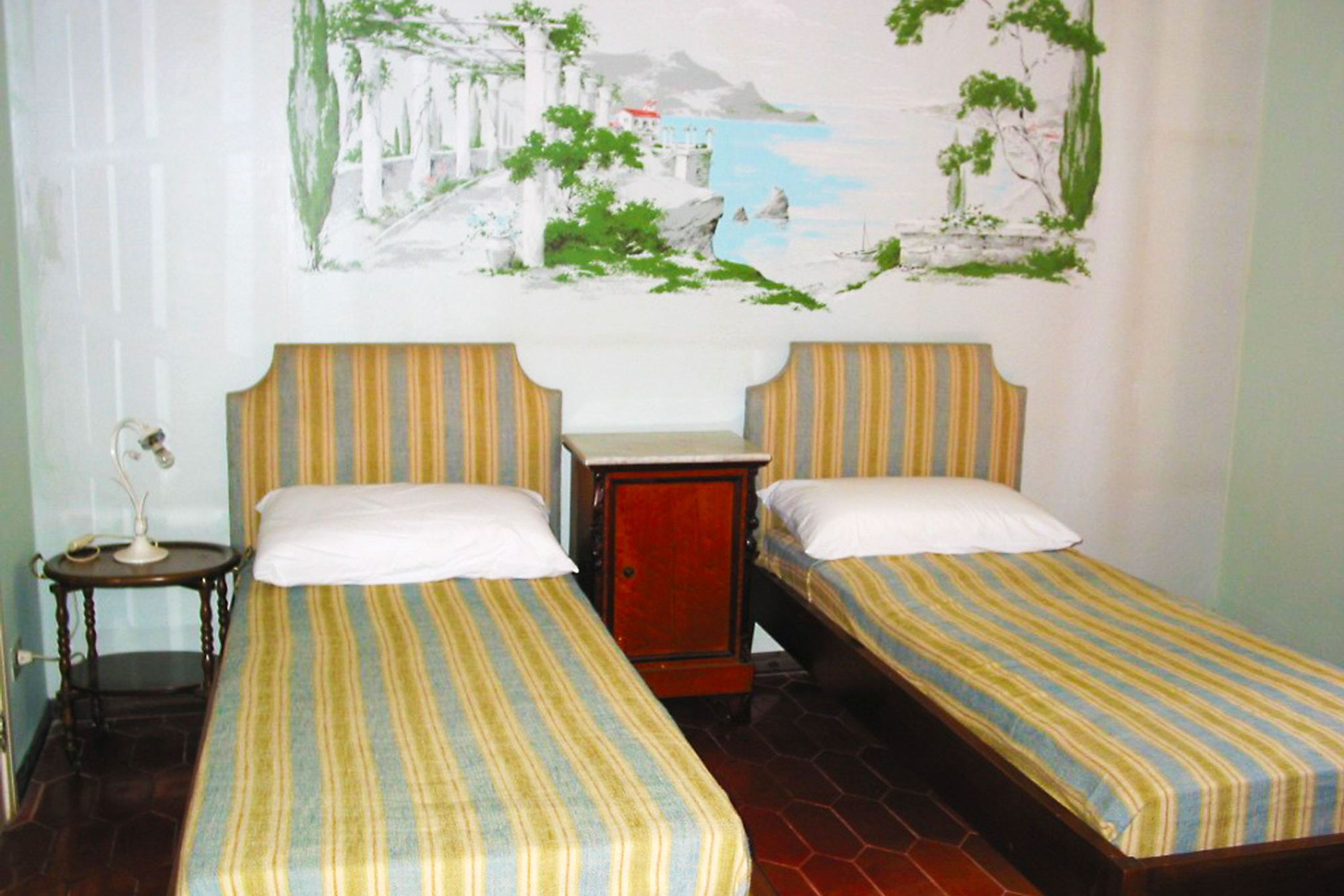 A room with two beds, each with striped bedding, a small wooden cabinet, and a wall painting.