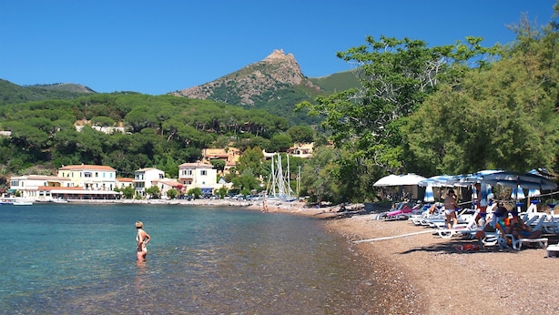 A beach with clear water, a sandy shore, and buildings in the background.