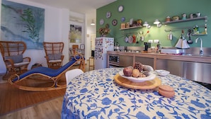 A living room with a blue floral sofa, wicker chairs, a dining table with a fruit centerpiece, and a kitchen with green walls and various kitchen items. - Appartamento Le Cannucce al Mare di Castiglione (Castiglione della Pescaia)