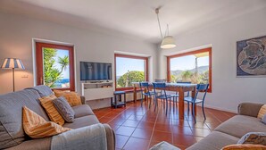 A spacious living area with a sofa, dining table, and chairs, large windows with a view, and a television.