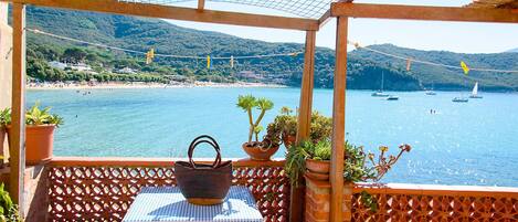A terrace with a table and chairs, overlooking a coastal area with boats and greenery.