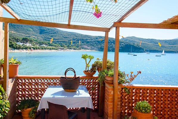 A terrace with a table and chairs, overlooking a coastal area with boats and greenery.