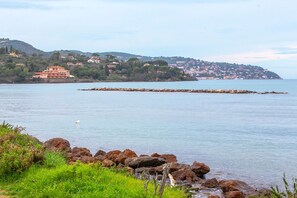 A coastal view with a rocky shoreline, a large building with multiple stories, and a distant hill with scattered houses.