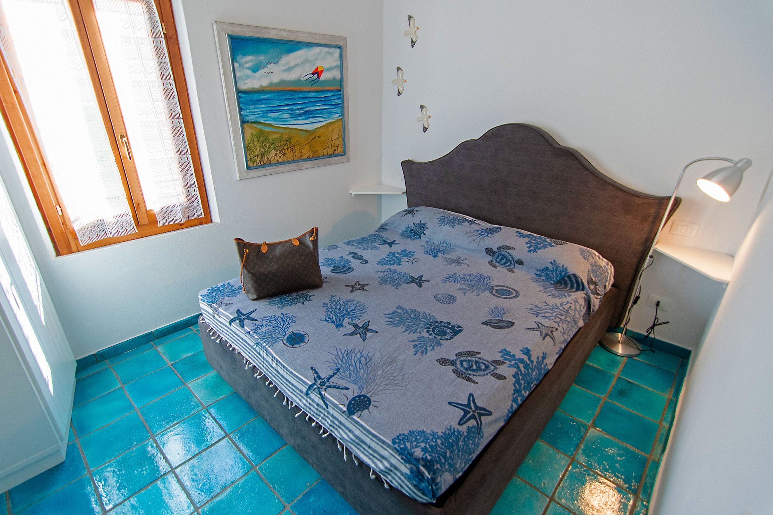 A bedroom with a bed, a window with curtains, a painting of a beach scene, and a tiled floor.