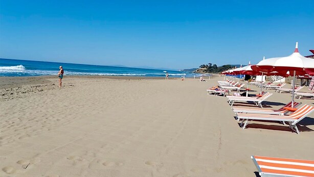 Une plage bordée de rangées de chaises longues et de parasols, un ciel dégagé et quelques personnes visibles au loin.