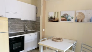 Une cuisine compacte avec des meubles blancs, un four encastré et un évier. On y trouve une table à manger avec quatre chaises et un panier en osier posé dessus.