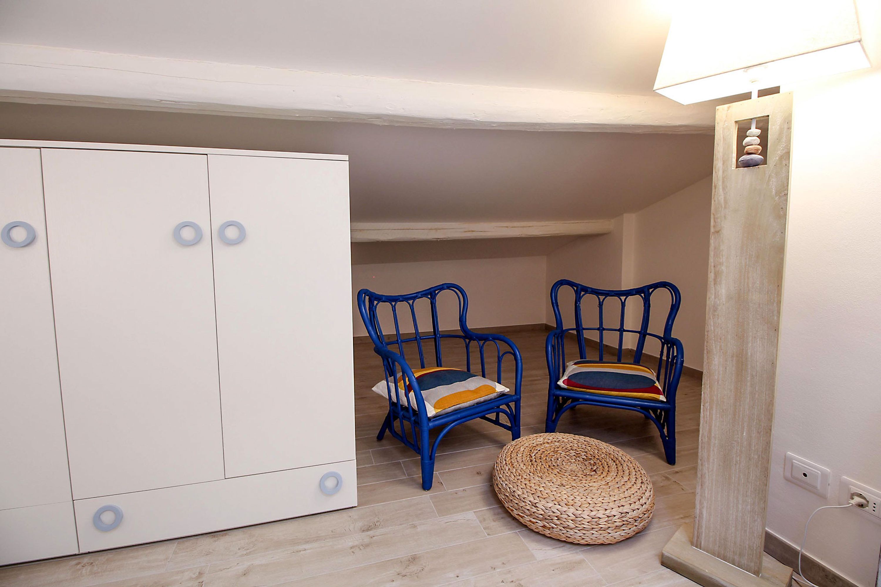A room with a wardrobe, two blue chairs with cushions, a woven pouf, and a lamp.