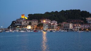 A coastal town at dusk with buildings along the waterfront, boats docked, and a prominent tower on a hill.
