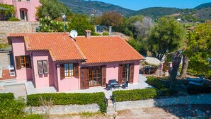 A pink villa with a red-tiled roof, surrounded by greenery and hills.