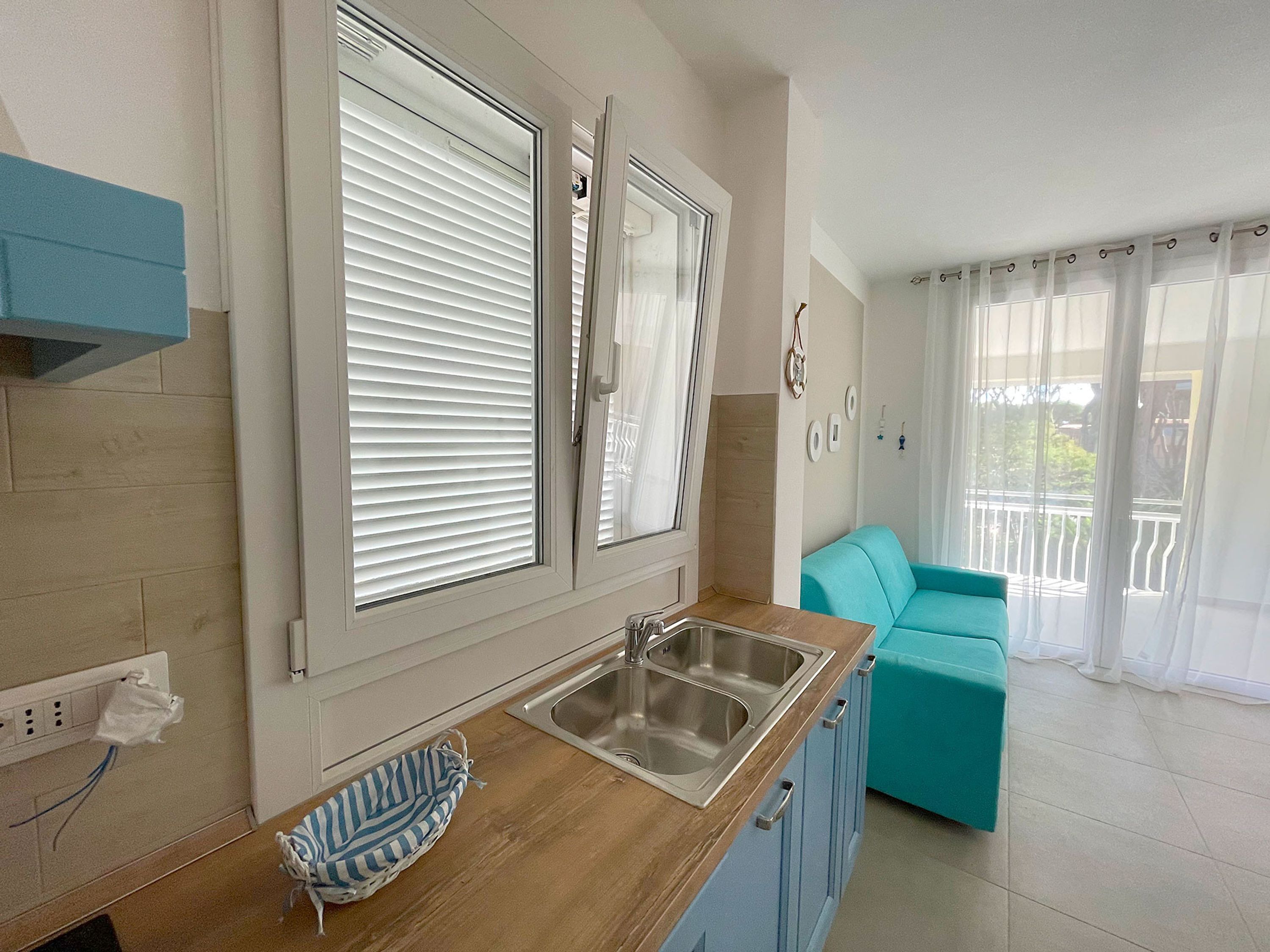 Una cocina con un lavamanos, una ventana con persianas, un sofá color turquesa y un balcón con barandilla.