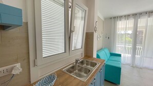 A kitchen with a sink, a window with blinds, a teal sofa, and a balcony with a railing.
