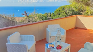 Un balcon agrémenté de meubles en osier blanc, une petite table avec un livre et des fruits, et une vue sur la mer.
