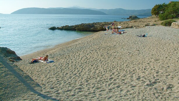 Une plage de sable où des gens se prélassent au soleil, avec une vue dégagée sur la mer et les montagnes lointaines.