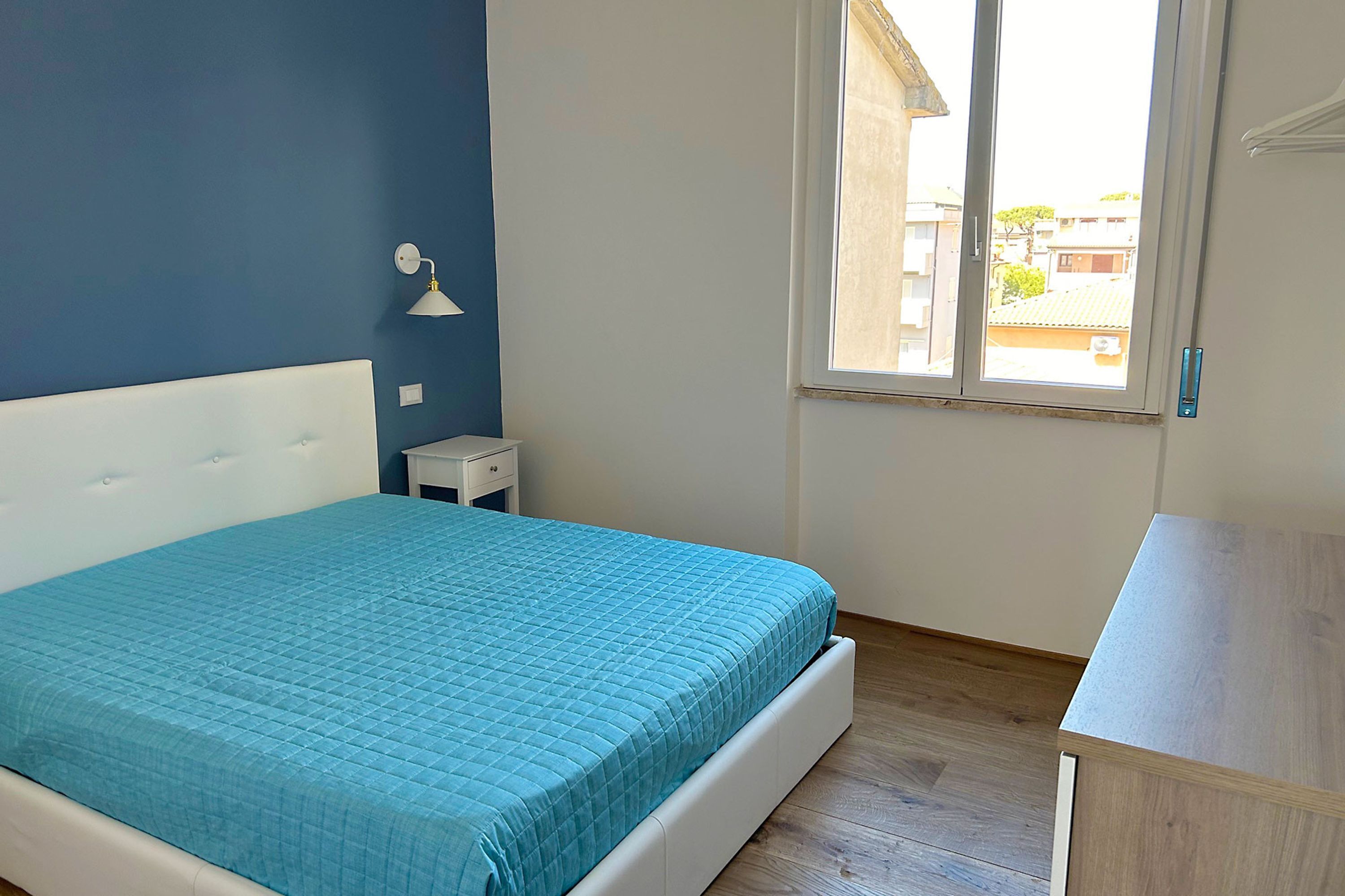 A bedroom with a bed, a nightstand, a desk, a window, and a view of the outside.